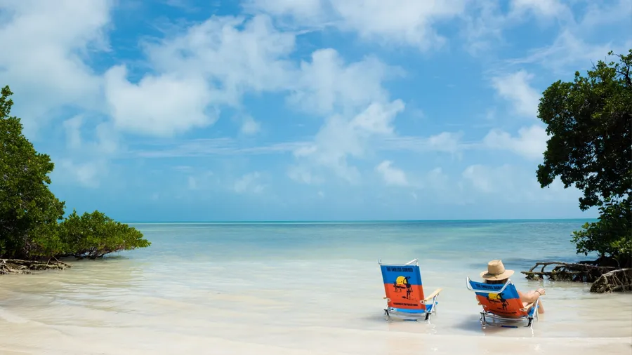 Person relaxing in beach chair beside empty chair on calm tropical ocean shoreline with blue sky and copy space