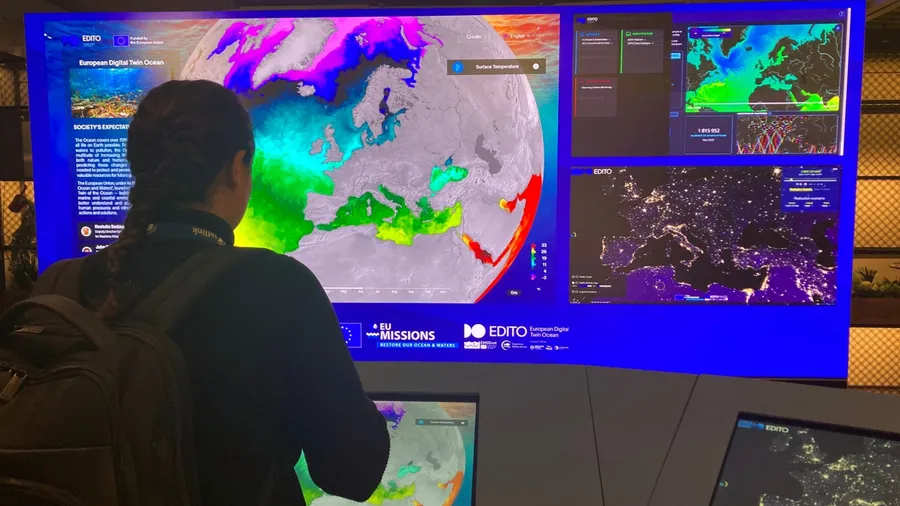 Brussels, Belgium-26 November 2025. A person observing a large digital screen displaying a map of the Earth with colored overlays representing oceanic data, part of the European Digital Twin Ocean