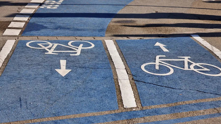 Urban bicycle lane with blue pavement and white directional bike symbols. Divided for two-way cycling traffic, marked with arrows and dashed lines. Promotes safe and organized cycling infrastructure.