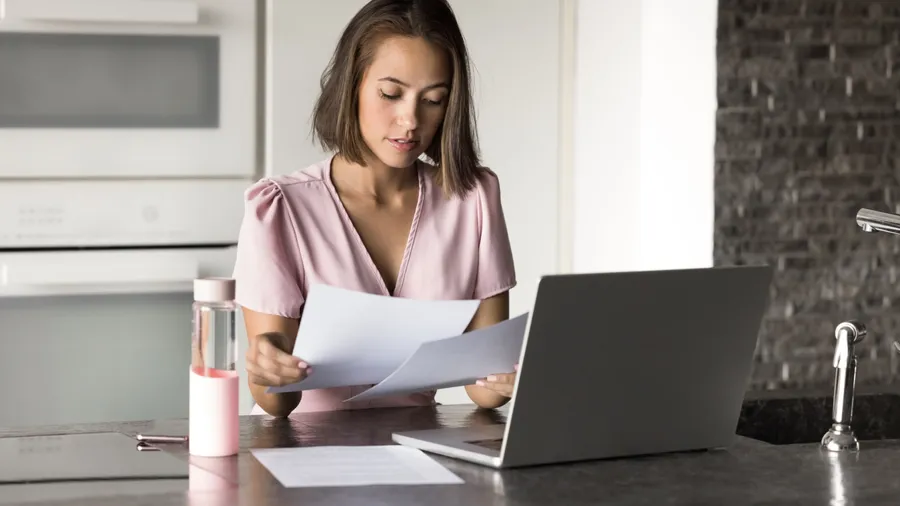 Young 25s woman sits at kitchen counter with laptop, focused on reviewing important documents, working on task, reviewing reports, contracts, or assignments. Teleworking, freelance, budget management