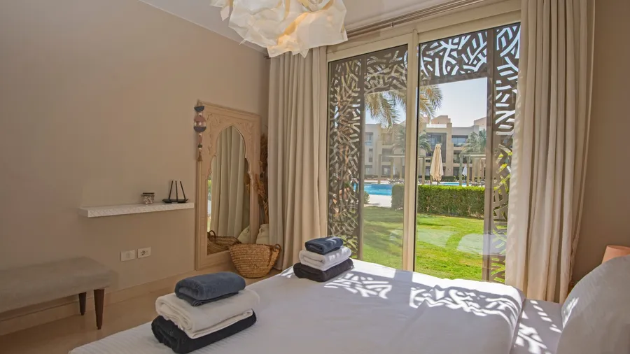 Bedroom with white linens, nautical decor, patterned screens, garden view, pool, palm trees, and sunlight.