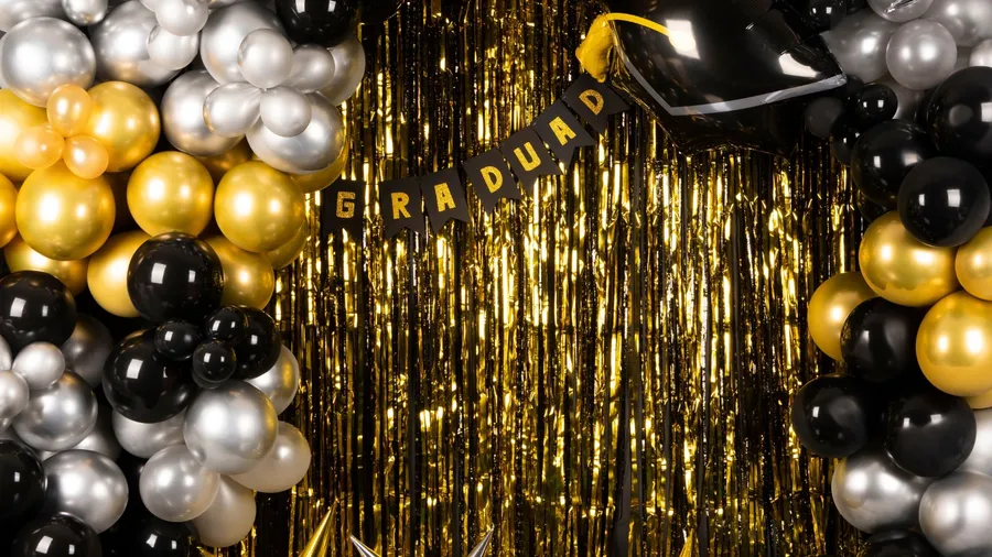 A stunning grad party decor with gold streamers backdrop and balloon arch with black, gold, and silver balloons.