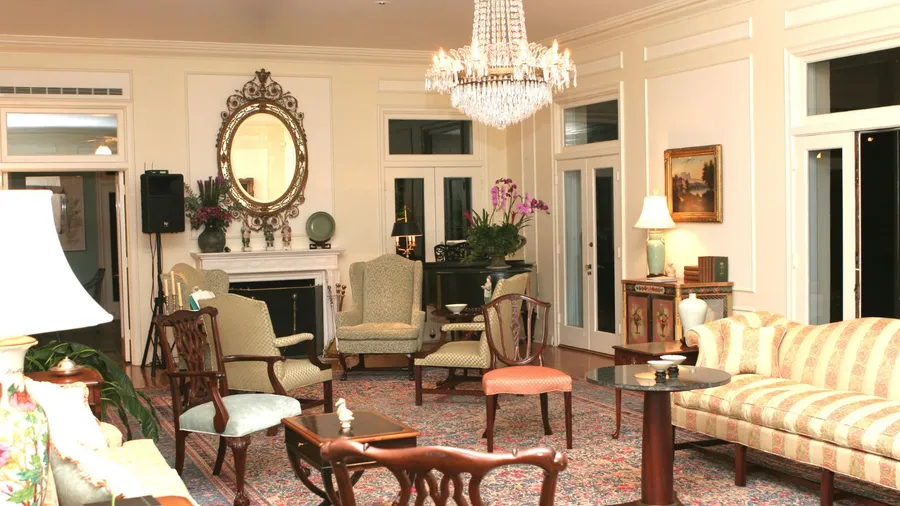 A formal living room with a grand chandelier hanging from the ceiling, an ornate gold mirror , and various antique-style furniture including a striped sofa, armchairs, and wooden tables, with French doors leading outside.