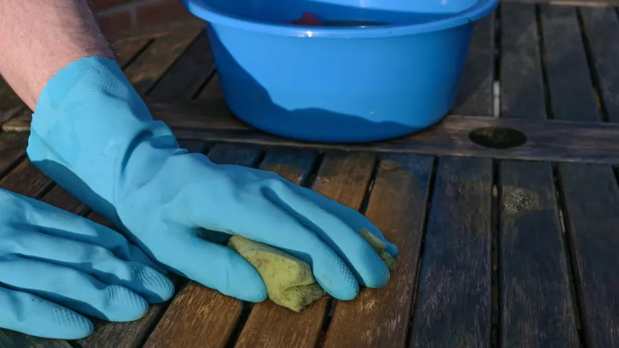 A person wearing blue gloves scrubbing the wooden surface using a sponge with a blue basket near him.