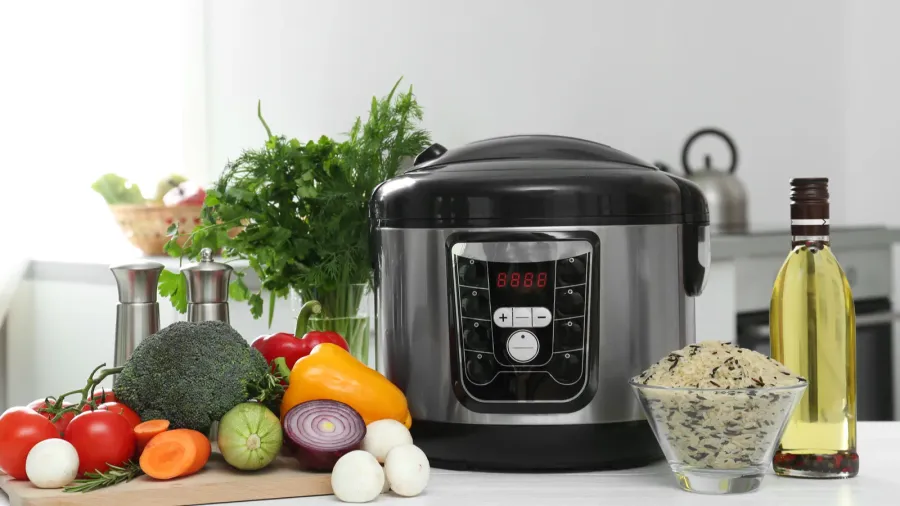 A rice cooker, fresh produce, rice in a bowl, herbs, and a cooking oil bottle on a kitchen countertop.