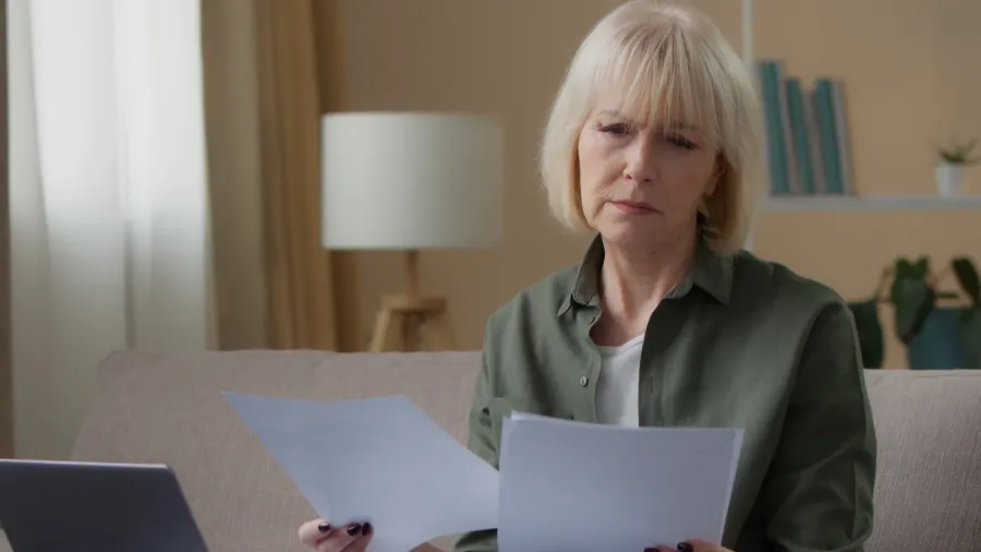 serious caucasian old woman with papers documents looking laptop confused