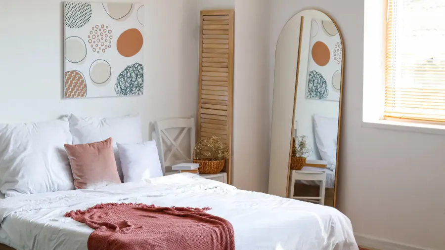 A stylish modern bedroom interior with cozy bed, abstract artwork, and full-length mirror near a window.