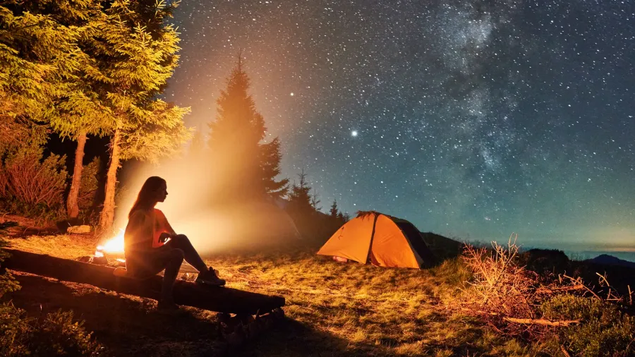 young woman hiker sitting on bench near bonfire under magical